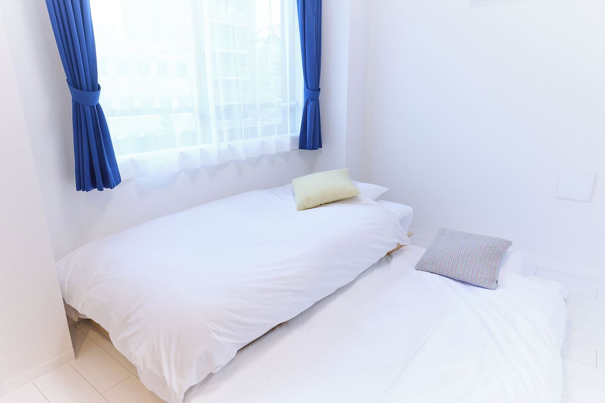 A single bed with white bedding, a yellow pillow, and a patterned pillow. Blue curtains are tied at the top of the window.
