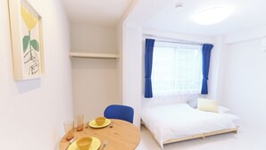A hotel room with a bed, a dining table with yellow plates and cutlery, a blue chair, and a window with blue curtains. - Zenith Nishinakanobu (Tokyo)