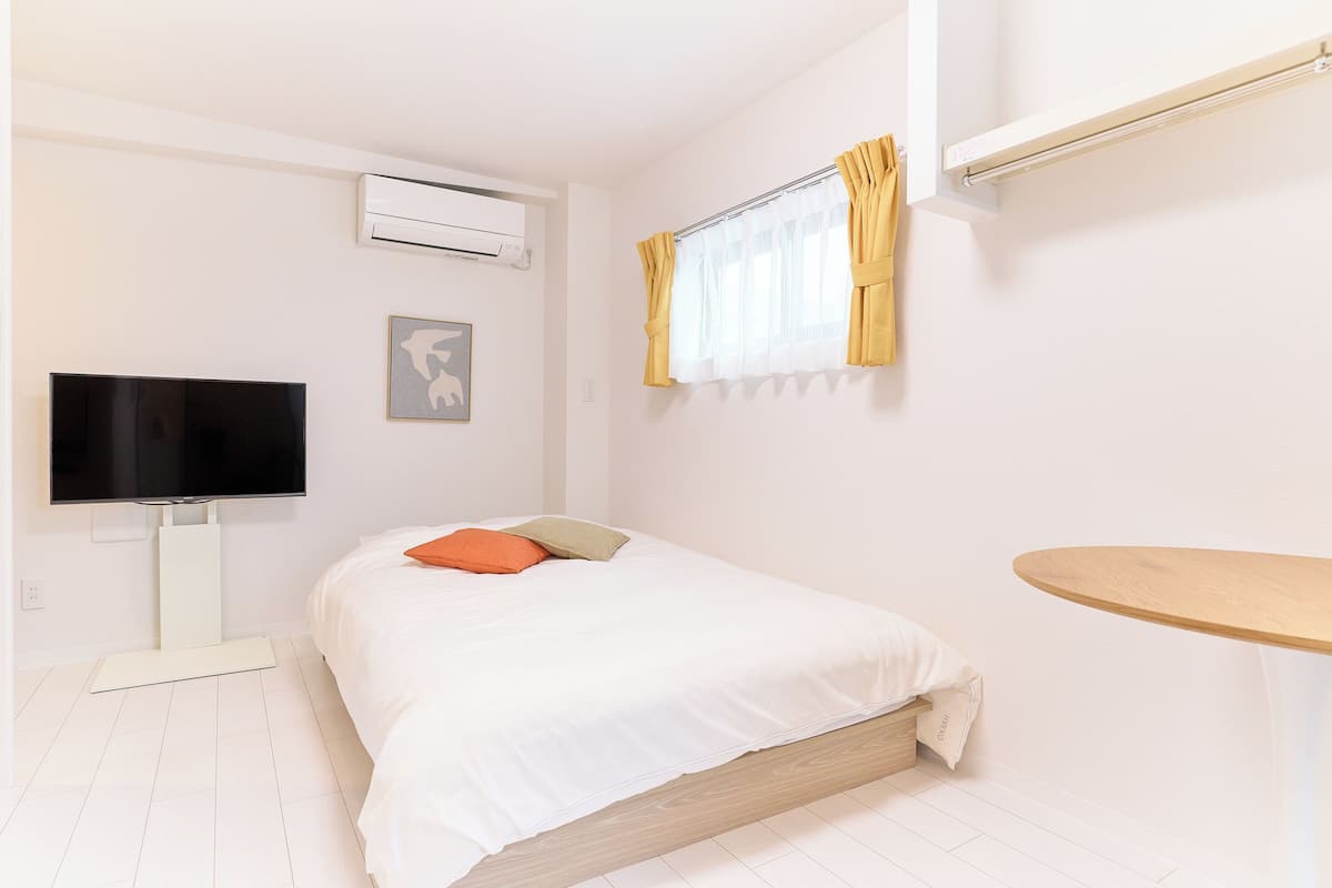 A bedroom with a bed, a TV, an air conditioner, a window with curtains, and a small wooden table.