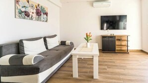 A modern living room with a grey sofa, a white coffee table, a painting on the wall, a flat-screen TV, and a wooden cabinet. - Apartment 3 (Novalja)