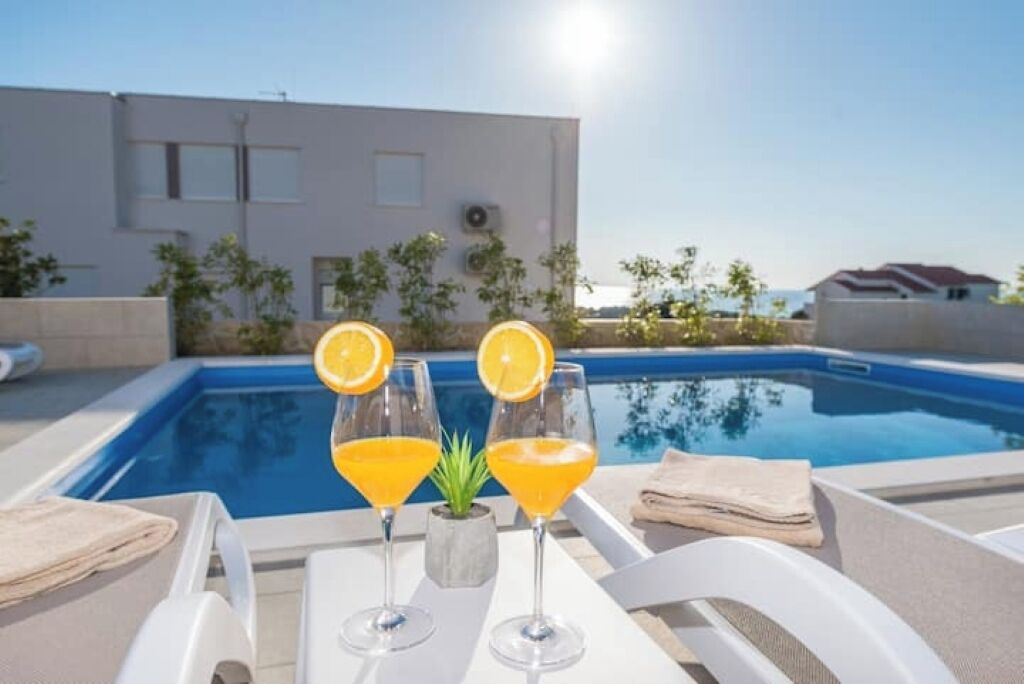 A poolside area with a clear view of a building in the background, a glass table with two drinks and a small plant, and a towel on the chair.