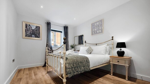 Un dormitorio con piso de madera, una cama con ropa blanca, una mesita de noche con lámpara y un ventanal con cortinas.