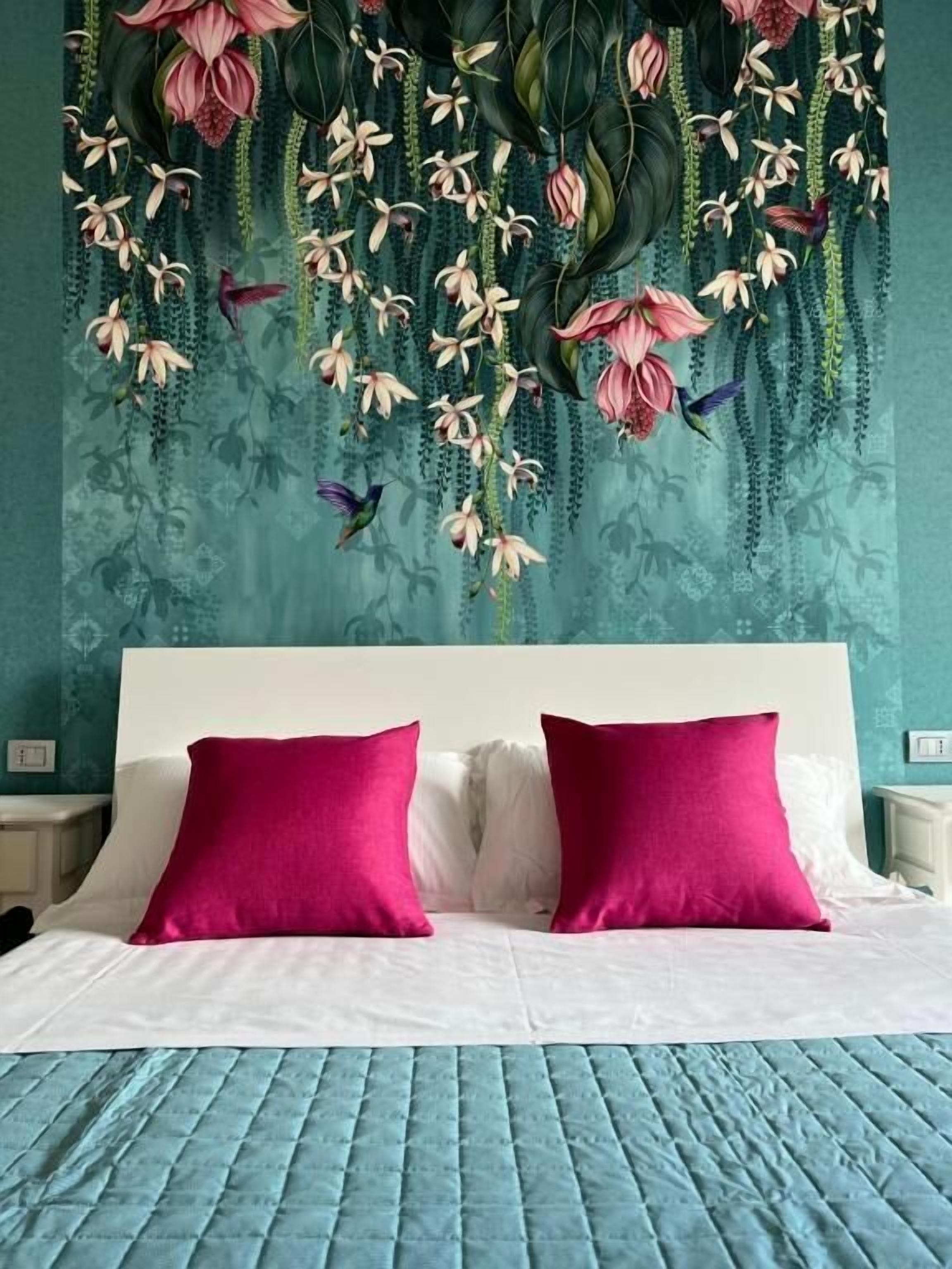 Un lit avec une literie blanche et deux coussins rose vif, un mur de couleur turquoise orné d’un papier peint à motifs floraux, et une composition florale suspendue au-dessus du lit.