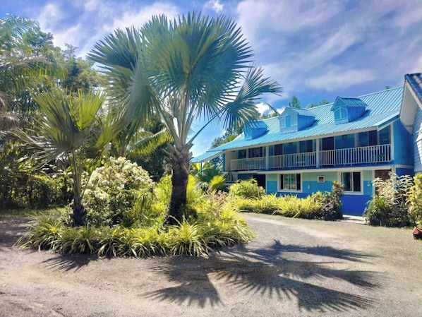 Un edificio de dos pisos con techo azul y paredes blancas, rodeado de exuberante vegetación tropical.