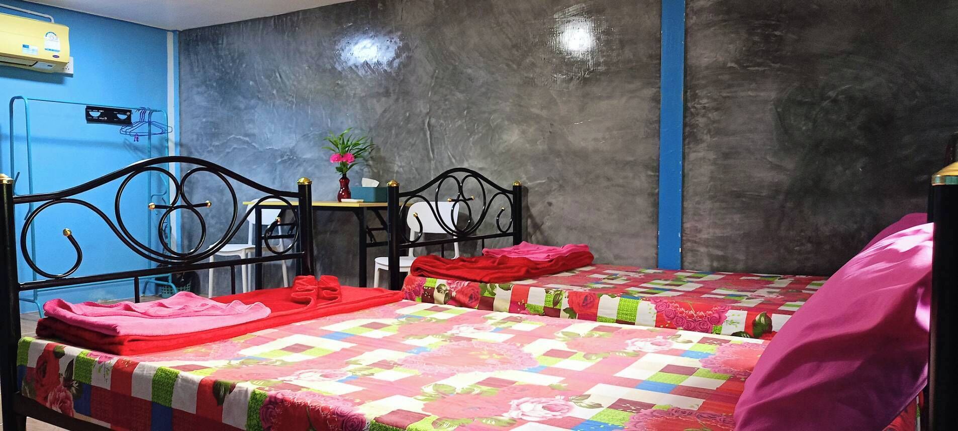 A hotel room with two beds, each with a floral quilt, a small table with a vase, and a blue wall.