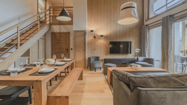 A spacious living area with a wooden staircase, a dining table set for four, a comfortable sofa, and a flat-screen TV mounted on the wall. - Apartment in The Residence L' Éclat des  Véria (MEGEVE)