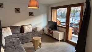 A modern living room with a sectional sofa, a television on a white cabinet, a wooden stool, and a large window offering a city view. - Les Marmottes N°302 - 2P7 (Les Deux Alpes)