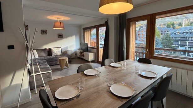 A modern dining area with a wooden table set for four, a sofa, a TV, and a view of buildings through large windows. - Les Marmottes N°302 - 2P7 (Les Deux Alpes)