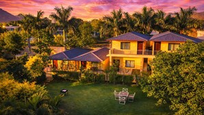 A two-story house with a balcony, surrounded by lush greenery and palm trees, during sunset. - Rambagh Retreat-Stay with  Swimming Pool (Udaipur)