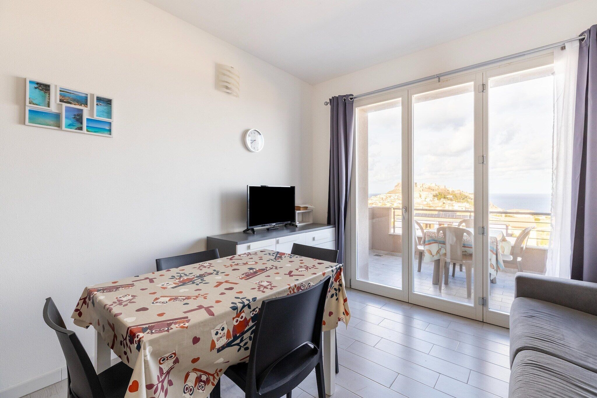 A room with a dining table, chairs, a television, a clock, and a balcony with a view.