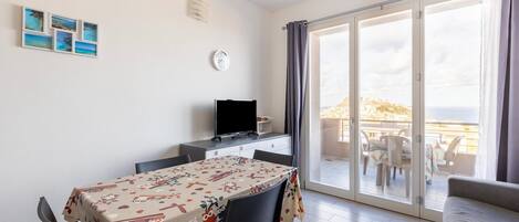 A room with a dining table, chairs, a television, a clock, and a balcony with a view.