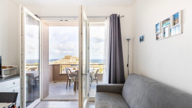 A room with a grey sofa, a sliding glass door leading to a balcony with a table and chairs, and a view of a coastal landscape. - Air conditioning, swimming pool and parking space (Castelsardo)