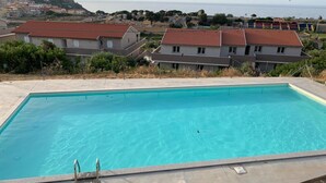 A rectangular swimming pool with a clear blue water surface, surrounded by a paved deck. In the background, there are houses with red-tiled roofs and a hilly landscape with a view of the sea. - Air conditioning, swimming pool and parking space (Castelsardo)