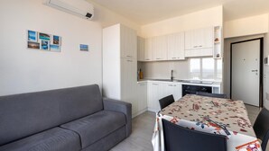 A compact living space with a grey sofa, a dining table with a patterned tablecloth, and a kitchenette with white cabinets. - Air conditioning, swimming pool and parking space (Castelsardo)