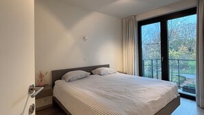 A modern bedroom with a large bed, a nightstand, a window with curtains, and a view of trees outside. - La Petite Foret Modern Flat Near Historic Bruges (Bruges)