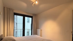 A bedroom with a large bed, a window with curtains, a radiator, and a ceiling light fixture. - La Petite Foret Modern Flat Near Historic Bruges (Bruges)