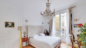 A bedroom with a large bed, a chandelier, a view of a building through a window, and a plant.