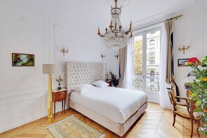 A bedroom with a large bed, a chandelier, a view of a building through a window, and a plant.