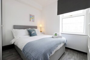 Un lit bien fait, avec une tête de lit grise, une couverture bleue et un oreiller orné d’un motif d’oiseau. On aperçoit une petite table de chevet sur laquelle se trouvent une lampe et une serviette. Une fenêtre donne sur un immeuble.