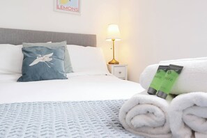 Un lit bien fait, avec un oreiller orné d’un motif d’oiseau, une lampe de chevet et une serviette roulée accompagnée de produits de toilette de la marque « pure pure ».
