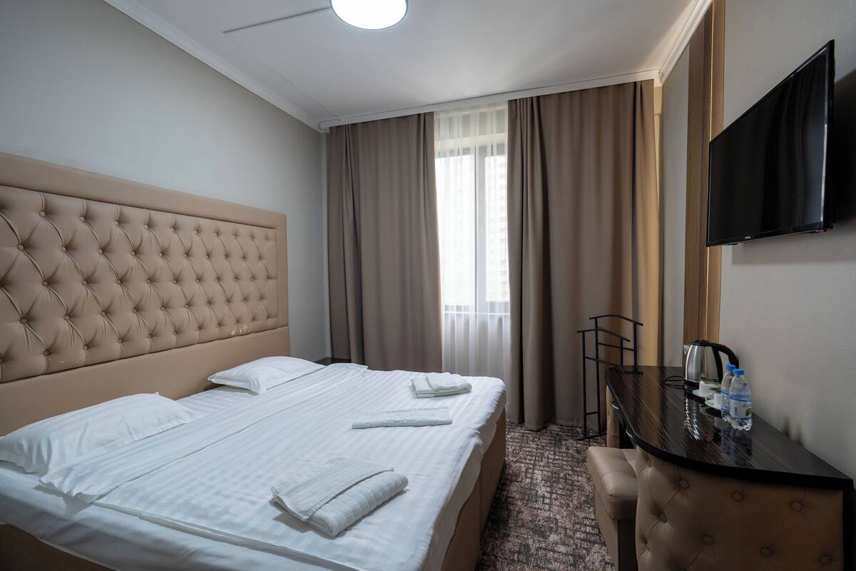 A hotel room with a large bed, a flat-screen TV, a desk with a chair, and a window with curtains.