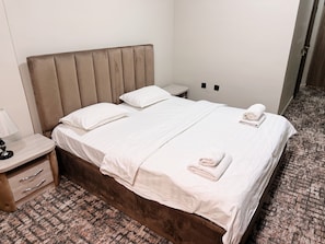 Een net opgemaakt bed met witte lakens, een houten nachtkastje met een lamp en een kamer met een gestreept tapijt.