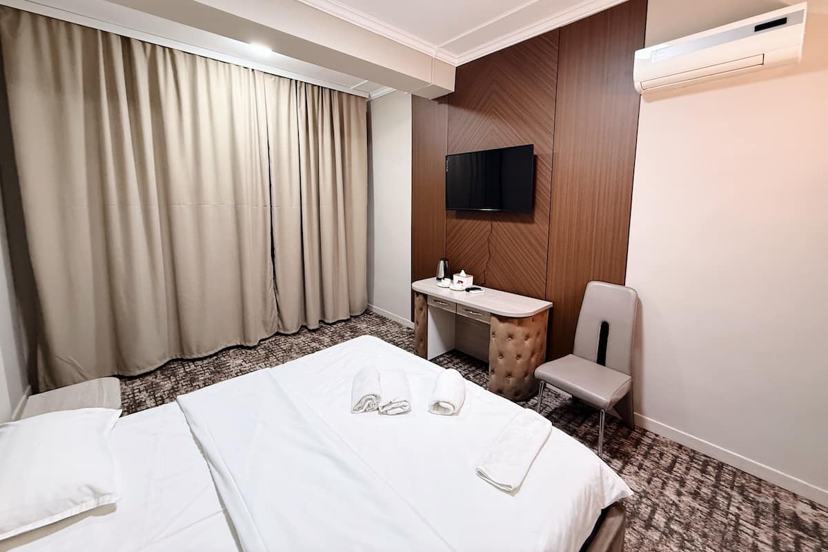A hotel room with a bed, a desk, a chair, a TV, and an air conditioning unit.