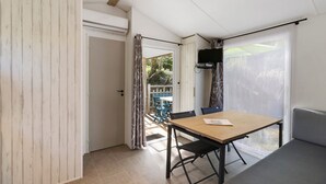 Une pièce comprenant une table en bois, deux chaises, un canapé, un téléviseur fixé au mur et une porte-fenêtre coulissante donnant sur un espace extérieur avec une table et des chaises.