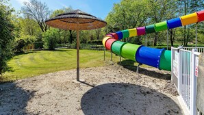 Un terrain de jeux avec un toboggan coloré et un parasol en chaume.