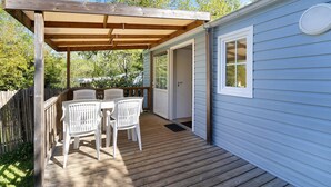 Une terrasse en bois, agrémentée de chaises blanches et d’une pergola, mène à un petit bâtiment doté d’une porte et d’une fenêtre.