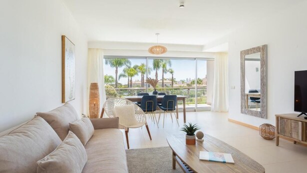 Ein modernes Wohnzimmer mit Sofa, Couchtisch und Fernseher. Ein Balkon bietet Blick auf Palmen und Gebäude.