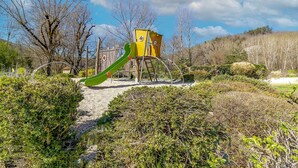 Un parque infantil con tobogán y estructuras para escalar, rodeado de árboles y arbustos.