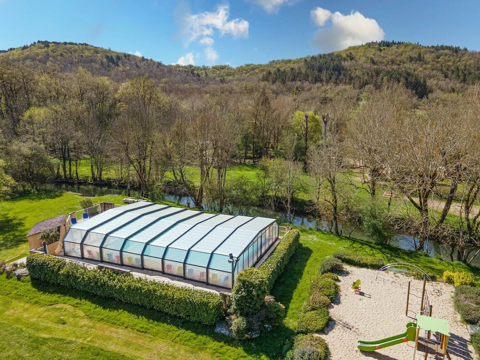 A large greenhouse structure with a glass roof, surrounded by well-maintained hedges and a sandy play area.