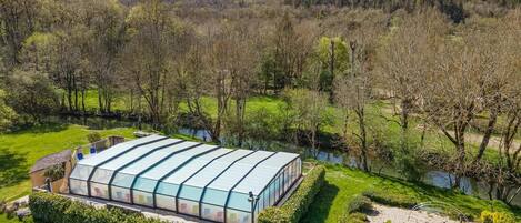 A large greenhouse structure with a glass roof, surrounded by well-maintained hedges and a sandy play area.