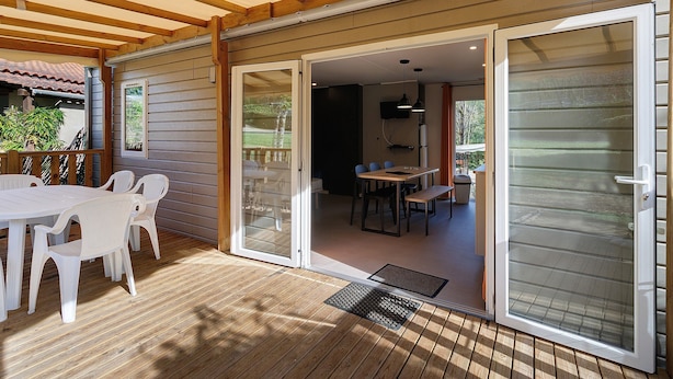 Een overdekt terras met een houten tafel en stoelen, een schuifdeur naar een eethoek binnen en een metalen ventilatie rooster op de vloer.