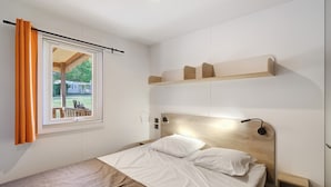 Een enkel bed met een houten hoofdbord, een nachtkastje, een raam met gordijnen en uitzicht op een huis en bomen.