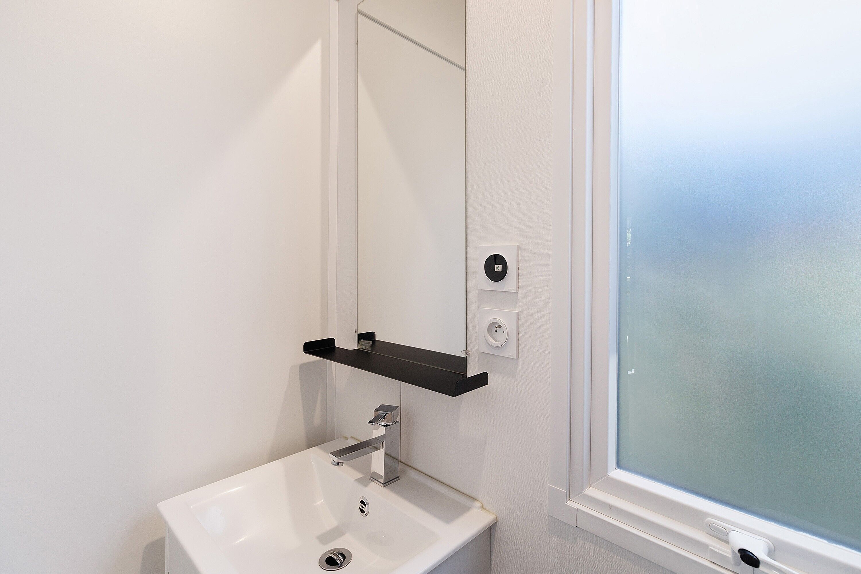 A bathroom with a white sink, a mirror, a black shelf, and a frosted window.