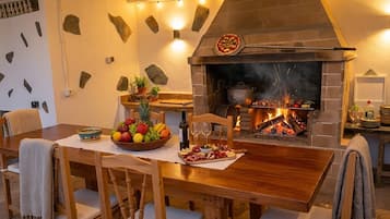 Een eetgedeelte met een bakstenen open haard, een pizzaoven, en een tafel gedekt met fruit, wijnglazen en een vleeswarenbord.