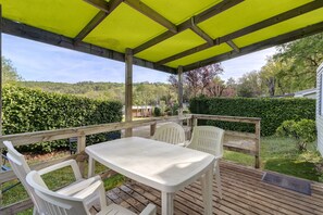 Une terrasse extérieure couverte, dotée d’une table et de chaises, entourée d’une plateforme en bois et offrant une vue sur un paysage verdoyant.