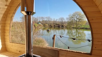 A wooden cabin with a large arched window overlooking a calm lake, a chimney, and a fishing rod.
