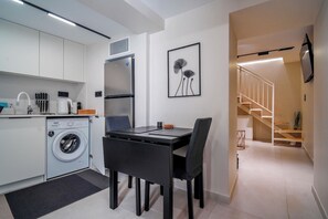 A modern kitchen with a washing machine, a dining area with a table and chairs, and a staircase leading to an upper level. - Pothos Luxury Maisonette (Iraklio)