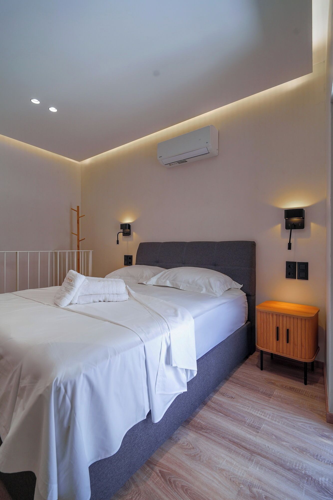 A modern bedroom with a large bed, bedside lamps, an air conditioning unit, and a wooden nightstand.