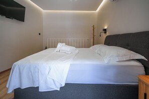 Cama bem arrumada com lençóis brancos e toalha dobrada, cabeceira estofada cinza e televisão de tela plana fixada na parede.