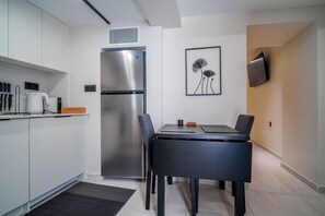 A compact kitchen with a built-in refrigerator, a dining area with a table and chairs, and a wall-mounted flat-screen TV. - Pothos Luxury Maisonette (Iraklio)