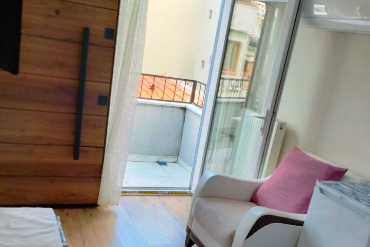 A hotel room with a bed, a sofa, a wooden wardrobe, a balcony with a railing, and a sliding glass door.