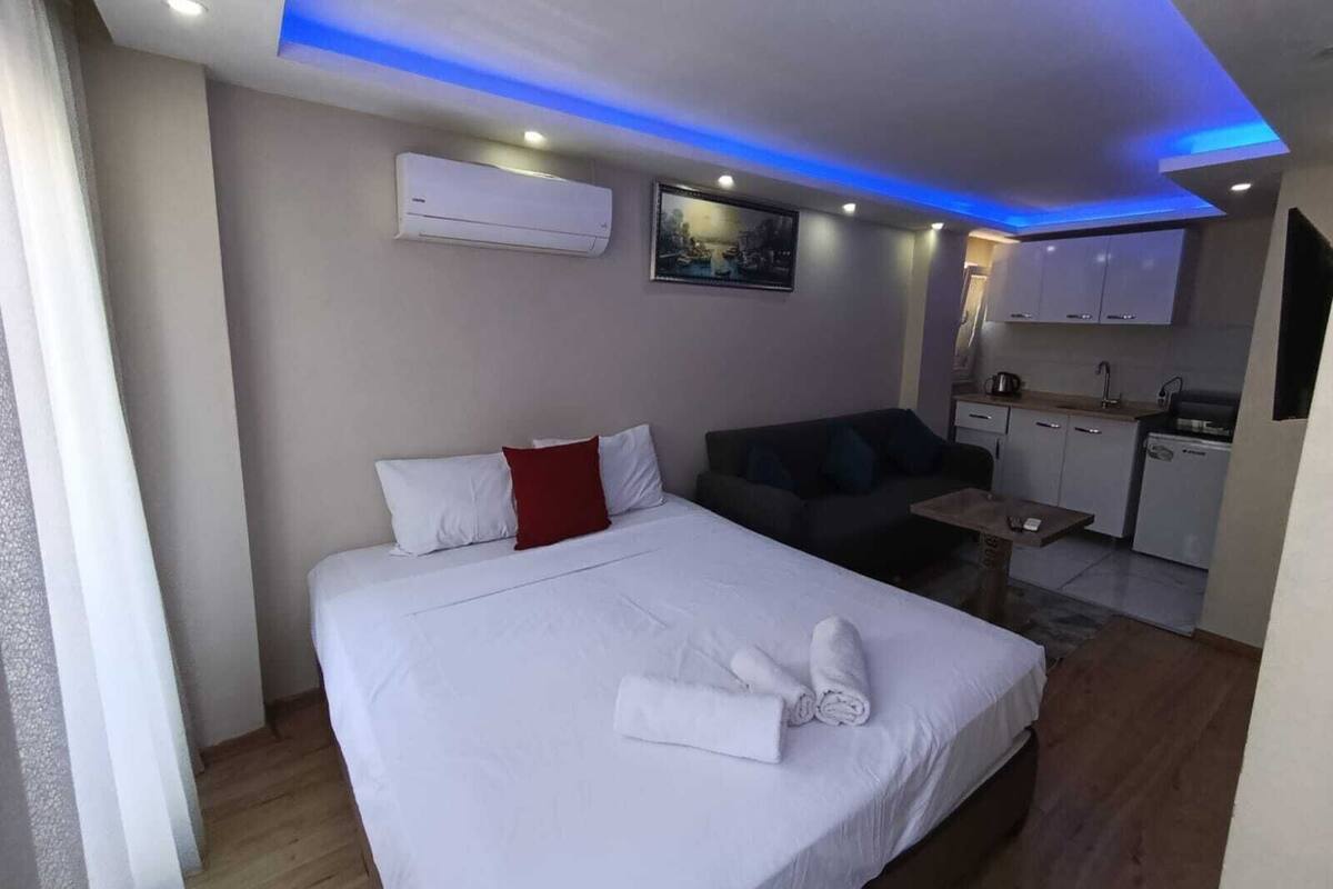 A hotel room with a bed, a sofa, a small table, an air conditioning unit, a painting, and a kitchenette.