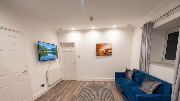 Ein Zimmer mit einem blauen Sofa, einem grauen Teppich, einem Fernseher an der Wand und einer Tür.