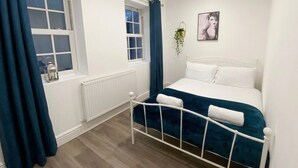 Ein Schlafzimmer mit Bett, Fenster mit Vorhängen, Heizkörper und einer Wandpflanze.
