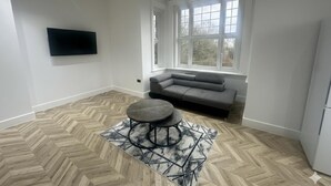 A modern living room with a herringbone floor, a grey sofa, a round coffee table, and a large window providing natural light.