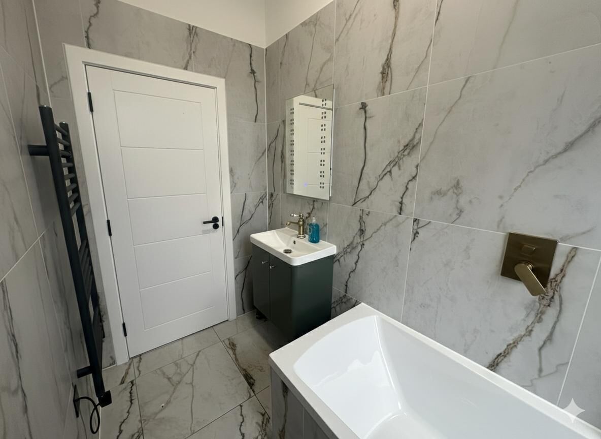 Une salle de bain avec une baignoire blanche, un lavabo avec un miroir au-dessus, et une porte. Les murs et le sol sont recouverts d'un motif imitant le marbre.